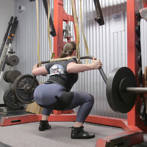 A person in workout clothes uses a safety squat bar with weight plates at the gym, surrounded by various equipment like weight racks and a notice. Product displayed: Spud Inc Sled Pulling Belt.