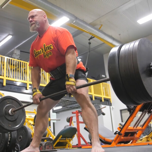 A bald man with a white beard deadlifts a heavily loaded barbell at a gym, wearing an orange t-shirt, black shorts, and Spud Inc Sled Pulling Belt. The gym features various workout machines and yellow railings.