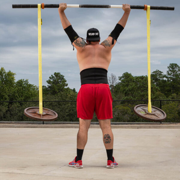 A person wearing a black cap, wristbands, red shorts, and sneakers is lifting a barbell with large weights attached by Spud Inc Swing Set Straps outdoors. The sky is cloudy and trees are visible in the background.