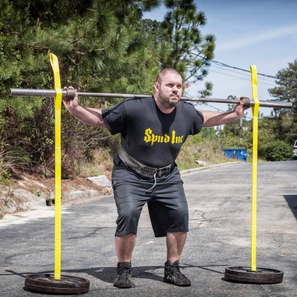 A man is squatting outdoors with a barbell secured by the Spud Inc Swing Set Straps. Hes wearing a black Spud Inc T-shirt, shorts, and sneakers. Trees and a blue dumpster can be seen in the background.