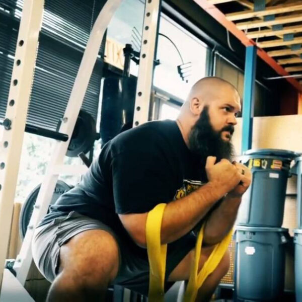 A bearded man performs a squat in a gym with Spud Inc Zercher Straps. He focuses intently, wearing a black t-shirt and gray shorts. Gym equipment and sunlight from a window set the background scene.