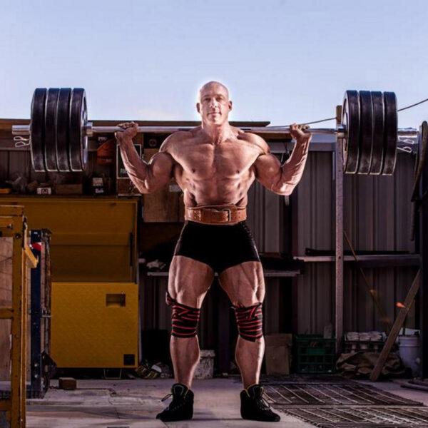A muscular man performs a heavy barbell squat outside, wearing shorts and the Sting Eco 4 Leather Belt. He has knee wraps and a determined expression, surrounded by gym equipment and a metal structure, all under a clear sky.