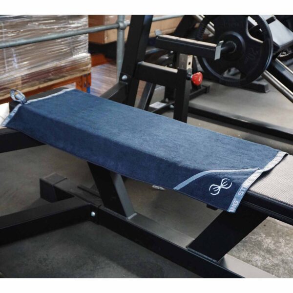 In a fitness center, a Sting Microfibre Gym Towel in blue is neatly placed on a black weight bench. Weight plates and a barbell can be seen behind the bench. The towel features a small logo in one corner.