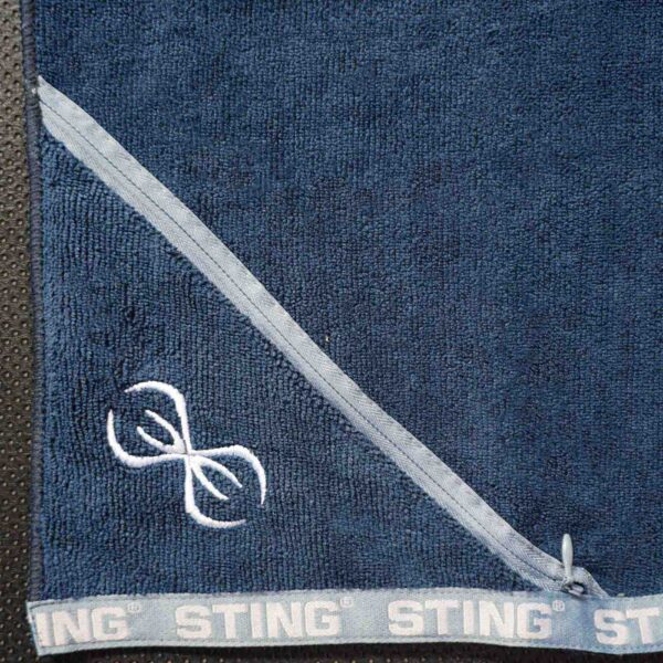 A close-up of the Sting Microfibre Gym Towel in dark blue, featuring a diagonal light blue stripe and a white embroidered logo with two stylized curved lines. The bottom edge repeats the word STING along the border.