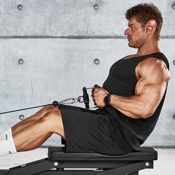 A muscular man with pointed ears is exercising with the ATX® Cable Row Handle Blackline. He is wearing a black tank top, black shorts, and white socks. The background shows a concrete wall adorned with circular patterns.
