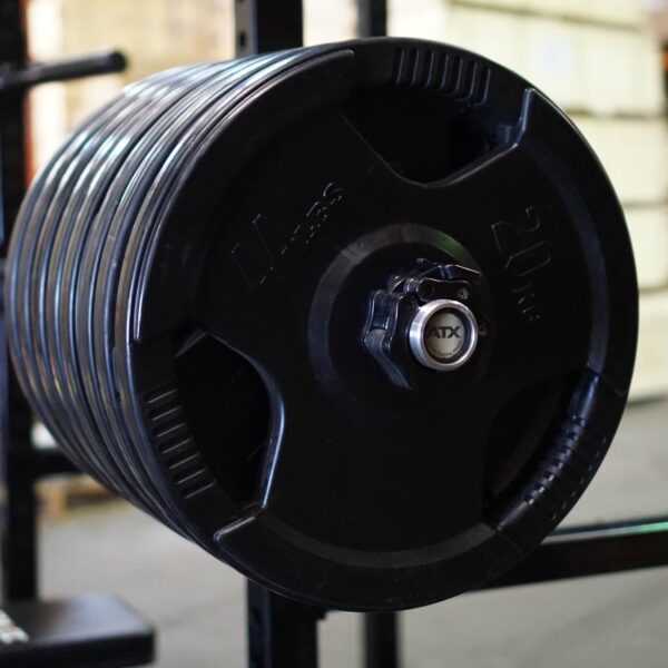 A close-up of the ATX® Olympic Power Bar Chrome loaded with several black weight plates, each labeled 20 kg, stacked on a gym rack. One plate is in focus, while the gym equipment is softly blurred in the background.