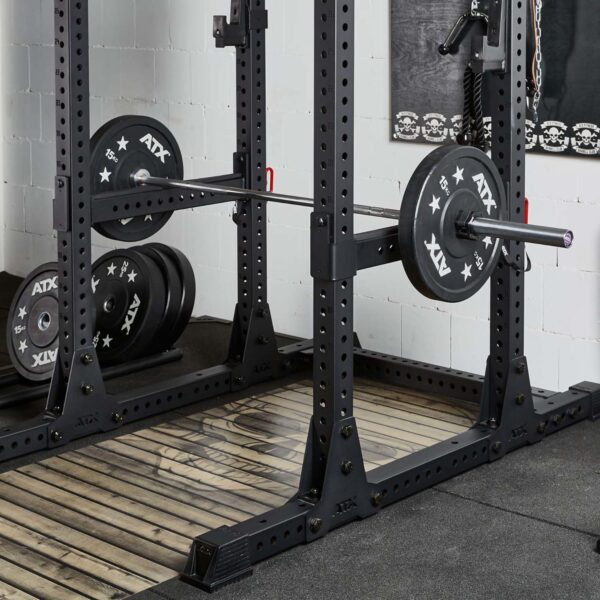 A weightlifting rack with a loaded barbell featuring ATX branding on the plates. The setup includes ATX® Spotter Bars 75cm on a wooden platform, with extra plates stacked on the left. The area is surrounded by gym equipment and chain decor on the wall.