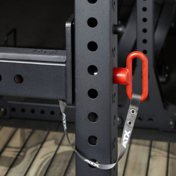 Close-up of a gym squat rack featuring the ATX® Spotter Bars 75cm with a red safety pin and strap attached to the black metal frame. Adjustable settings are available through multiple holes on the frame, which is set on a wooden floor.