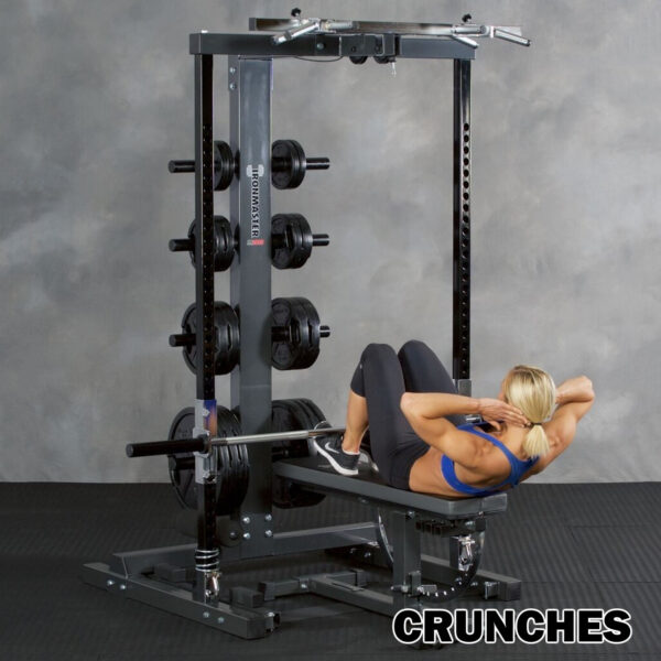 Wearing a blue sports bra and black leggings, a person performs crunches on an Ironmaster IM2000 Self-Spotting Smith Machine in a gym with various weights. At the bottom right, CRUNCHES is in bold.