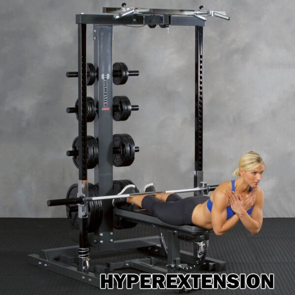 A person using the Ironmaster IM2000 Self-Spotting Smith Machine performs a hyperextension exercise, lying face down with their arms crossed and engaging lower back muscles. The machine features weights and cushioned leg support.