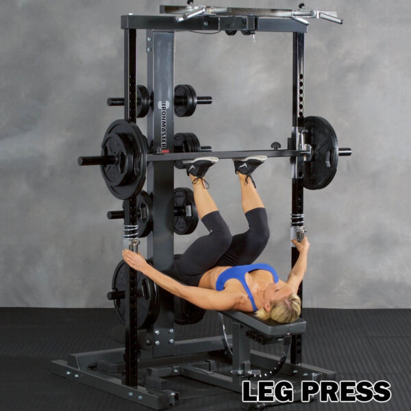 Clad in athletic wear, a person uses the Ironmaster IM2000 Self-Spotting Smith Machine to perform a vertical leg press, pushing a weighted platform with their feet while lying on a bench. Various weight plates surround them with LEG PRESS displayed at the bottom.