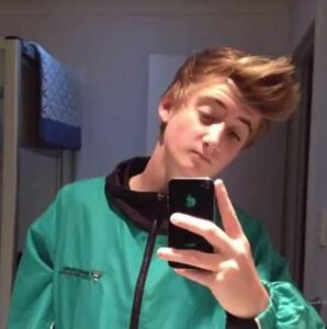 A person with styled, voluminous hair poses for a selfie in a mirror. They are wearing a green and black jacket and holding a smartphone. The background shows a towel on a rack.
