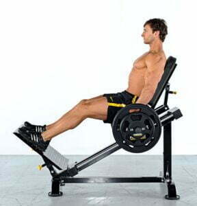A man in athletic shorts performs a leg press exercise on a machine. He is seated at an angle, pushing against weighted plates with his legs extended. The background is plain white.
