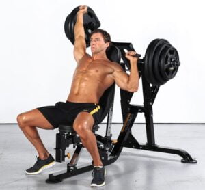 A muscular man is seated on a machine in the gym, performing a shoulder press with weight plates. He is shirtless, wearing black shorts with yellow stripes, and black sneakers. The background is plain white.