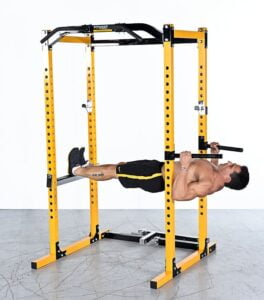 A man performs an inverted row using a bright yellow power rack in a gym setting. Hes gripping the bar with both hands, legs extended, and body parallel to the ground. The floor is gray, and the background is plain white.