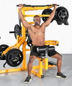 A person is seated on a yellow gym machine, holding a barbell overhead while smiling. They are wearing black shorts and black sneakers. Various weights are attached to the machine, and the background is plain white.