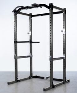 A black power rack for weightlifting standing on a gray floor. It features adjustable safety bars, a pull-up bar, and dip handles, all designed for strength training exercises.