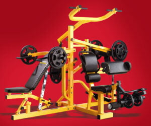 A yellow multi-station gym machine with various attachments, including a bench press, curl bar, and pull-up bar, set against a solid red background. The machine is equipped with multiple weight plates.