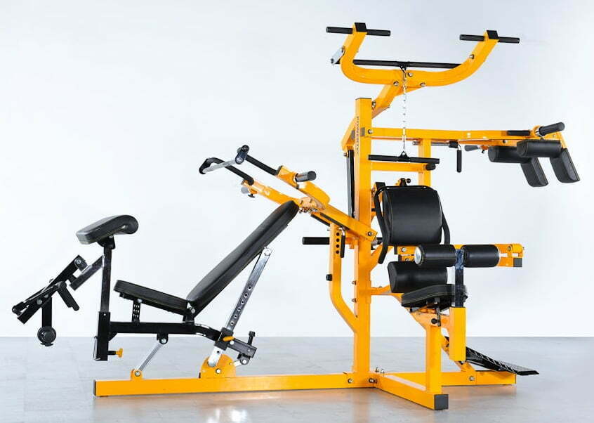 A multi-station home gym with a yellow frame and black cushioning against a plain background. It features an adjustable bench, pulleys, and various attachments for strength training exercises.