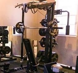 A gym with weightlifting equipment, including a squat rack with plates, a bench, and various machines. The room has light-colored walls and a window, creating a bright atmosphere.