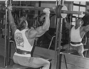 A person is performing a shoulder press with a barbell in a gym, sitting on a bench facing a mirror. They are wearing a tank top and shorts. The reflection shows their muscular build and focused expression.