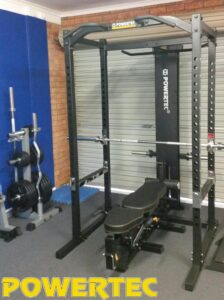A Powertec home gym setup featuring a power rack with a bench, barbell, and weight plates on a rack in the background. The equipment is placed in a garage with a rolled-up shutter door and a blue padded mat on the floor.