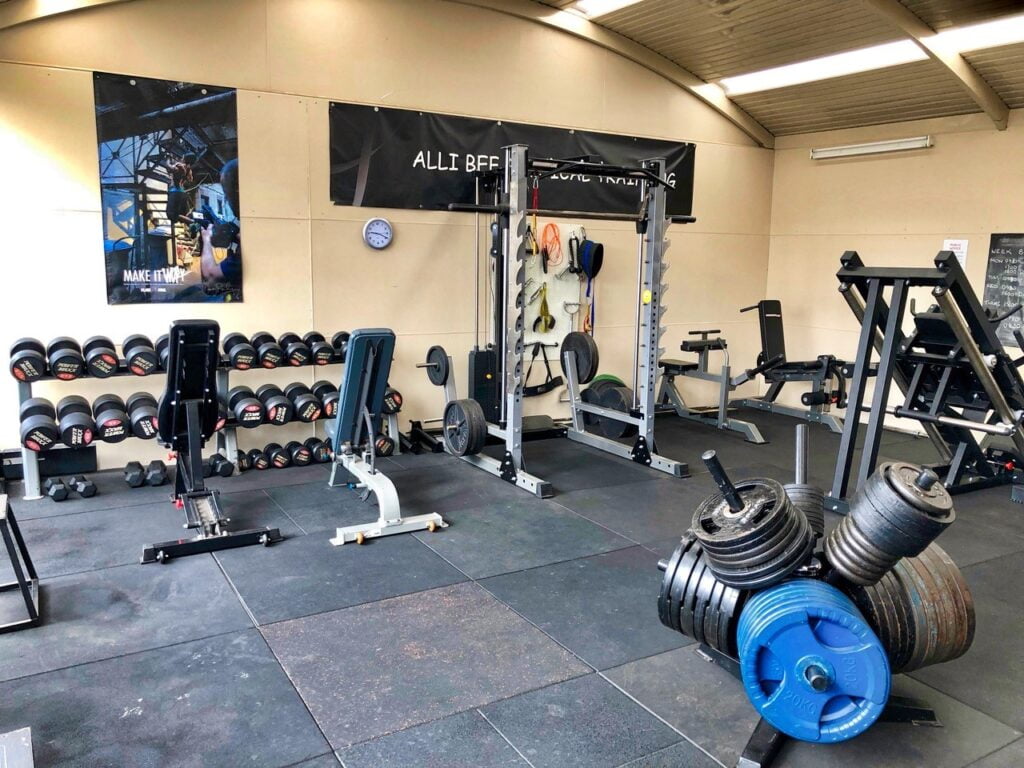 A spacious gym with weightlifting equipment. A rack of dumbbells is on the left, with benches and a squat rack in the center. Weight plates and a leg press machine are visible. Motivational posters hang on the walls.