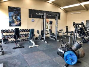 A spacious gym with weightlifting equipment. A rack of dumbbells is on the left, with benches and a squat rack in the center. Weight plates and a leg press machine are visible. Motivational posters hang on the walls.