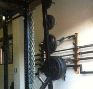 A home gym setup with a squat rack, weights, and lifting bars. Chains are hanging from the rack, and barbells are mounted on the wall. A jump rope is also visible in the background. The walls are painted white.