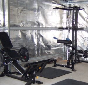 A home gym setup with a leg press machine on the left and a squat rack on the right. The walls are covered in metallic insulation, and the floor is padded with black exercise mats. The space is lit with natural light.
