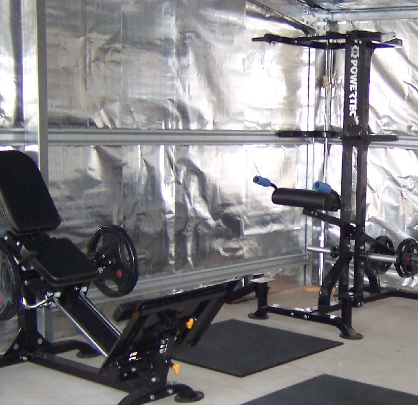 A home gym setup with a leg press machine on the left and a squat rack on the right. The walls are covered in metallic insulation, and the floor is padded with black exercise mats. The space is lit with natural light.