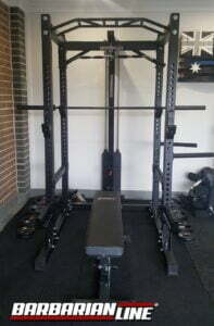 A home gym setup featuring a black Barbarian Line power rack with an adjustable bench. The equipment includes a pull-up bar, barbell, and weight plates. Theres an Australian flag on the wall in the background.