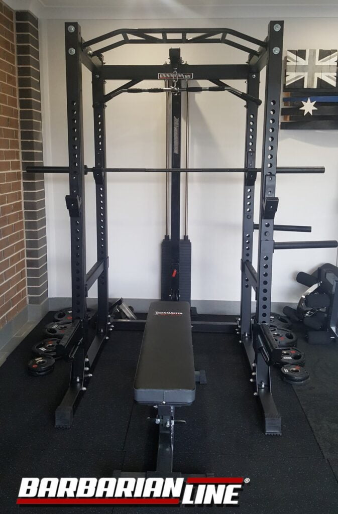A home gym setup featuring a black Barbarian Line power rack with an adjustable bench. The equipment includes a pull-up bar, barbell, and weight plates. Theres an Australian flag on the wall in the background.