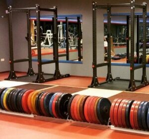 A gym with barbells and colorful weight plates neatly stacked on racks. There are squat racks in front of a large mirror, reflecting the gym equipment and surroundings. The floors are red, and the walls are blue and gray.