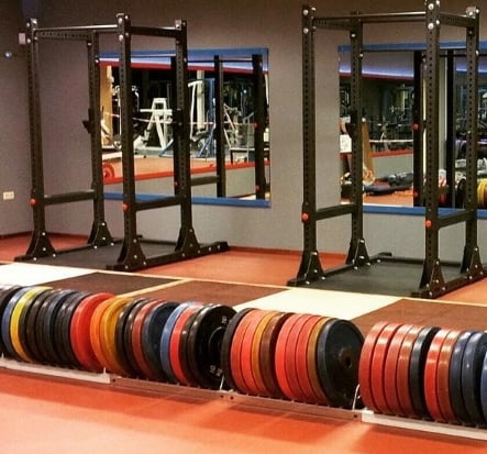 A gym with barbells and colorful weight plates neatly stacked on racks. There are squat racks in front of a large mirror, reflecting the gym equipment and surroundings. The floors are red, and the walls are blue and gray.