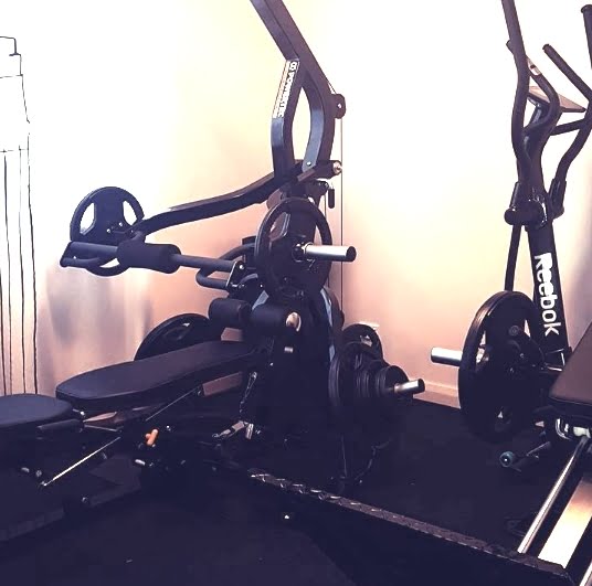 A small gym setup featuring weightlifting equipment, including a bench press with weights loaded onto the barbell. An exercise bike is positioned nearby, with a clear focus on fitness and strength training in a compact space.