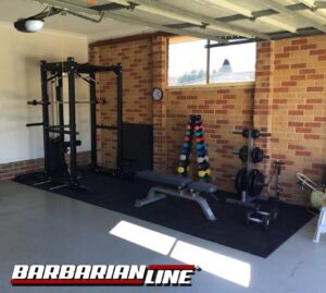 Home gym setup in a garage with a power rack, adjustable bench, and various free weights, including dumbbells and a weight tree. The space features brick walls and a small window. Logo in the bottom left corner reads Barbarian Line.