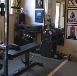 A home gym with various equipment, including a bench press, weight machine, and seated row machine. Walls are decorated with framed posters and a vintage Macintosh computer is placed on a shelf. A mannequin stands in a corner.