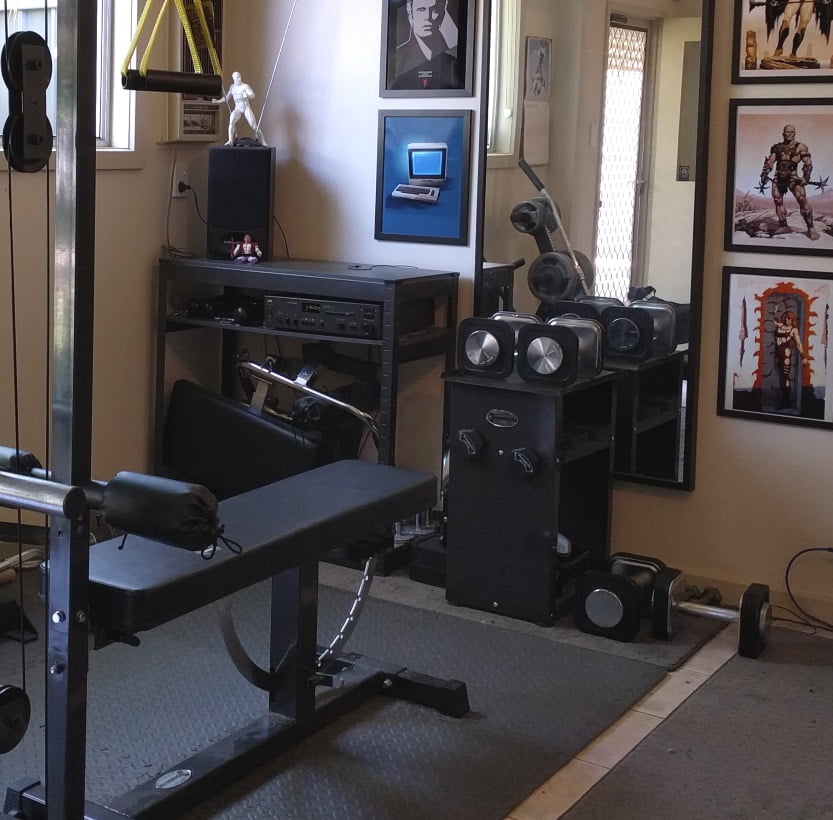 A home gym with various equipment, including a bench press, weight machine, and seated row machine. Walls are decorated with framed posters and a vintage Macintosh computer is placed on a shelf. A mannequin stands in a corner.