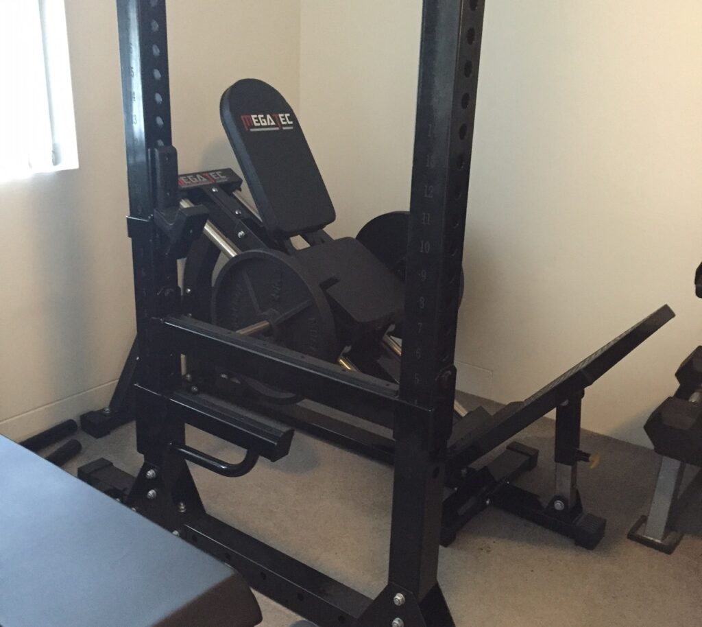 A gym room with exercise equipment including a squat rack, weight plates, and a bench. The room has a light-colored wall and a window on the left, allowing natural light to enter.