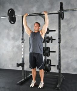 A man in a sleeveless shirt and shorts performs an overhead press with a barbell in a gym. He stands inside a power rack with weight plates on racks behind him, on a rubber mat floor.