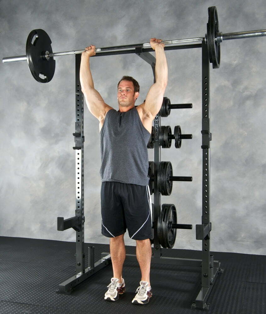 A man in a sleeveless shirt and shorts performs an overhead press with a barbell in a gym. He stands inside a power rack with weight plates on racks behind him, on a rubber mat floor.