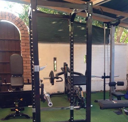 A home gym setup with a power rack, weight bench, and various gym equipment on a green floor. Its under a wooden roof with white walls and a brick archway entrance.