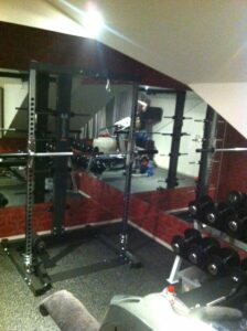 A home gym with a squat rack, dumbbells, and mirrored walls. Gym equipment is neatly arranged, including weights on a rack. A towel and water bottle are visible on a treadmill in the foreground.