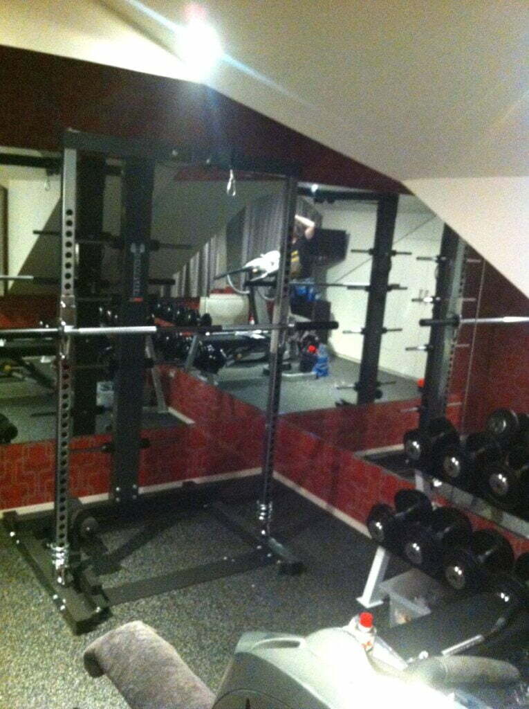 A home gym with a squat rack, dumbbells, and mirrored walls. Gym equipment is neatly arranged, including weights on a rack. A towel and water bottle are visible on a treadmill in the foreground.