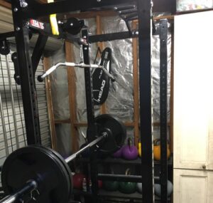 Home gym setup with a weightlifting rack, barbell, and lat pulldown attachment. Various colored kettlebells are stored on a shelf beneath the rack, and a tennis racket bag hangs on the frame. The room has a wooden and metal structure.