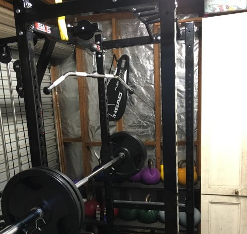 Home gym setup with a weightlifting rack, barbell, and lat pulldown attachment. Various colored kettlebells are stored on a shelf beneath the rack, and a tennis racket bag hangs on the frame. The room has a wooden and metal structure.