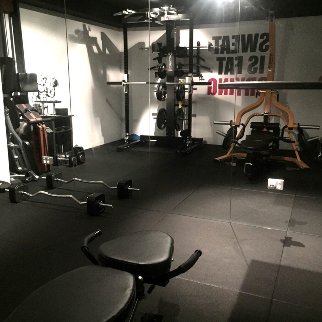 A dimly lit gym with weightlifting equipment, including benches, barbells, and a squat rack. A motivational quote on the wall reads, Sweat is fat crying. The floor is covered with black rubber mats.