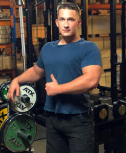 A man in a blue t-shirt and black pants stands in a gym, surrounded by weightlifting plates and equipment. He is giving a thumbs-up with a confident expression. The background is filled with industrial-style shelving.