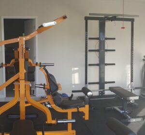 Home gym with various weightlifting equipment, including an orange multi-station machine, a squat rack with a pull-up bar, and benches. The room has a light color palette with some natural light coming in.